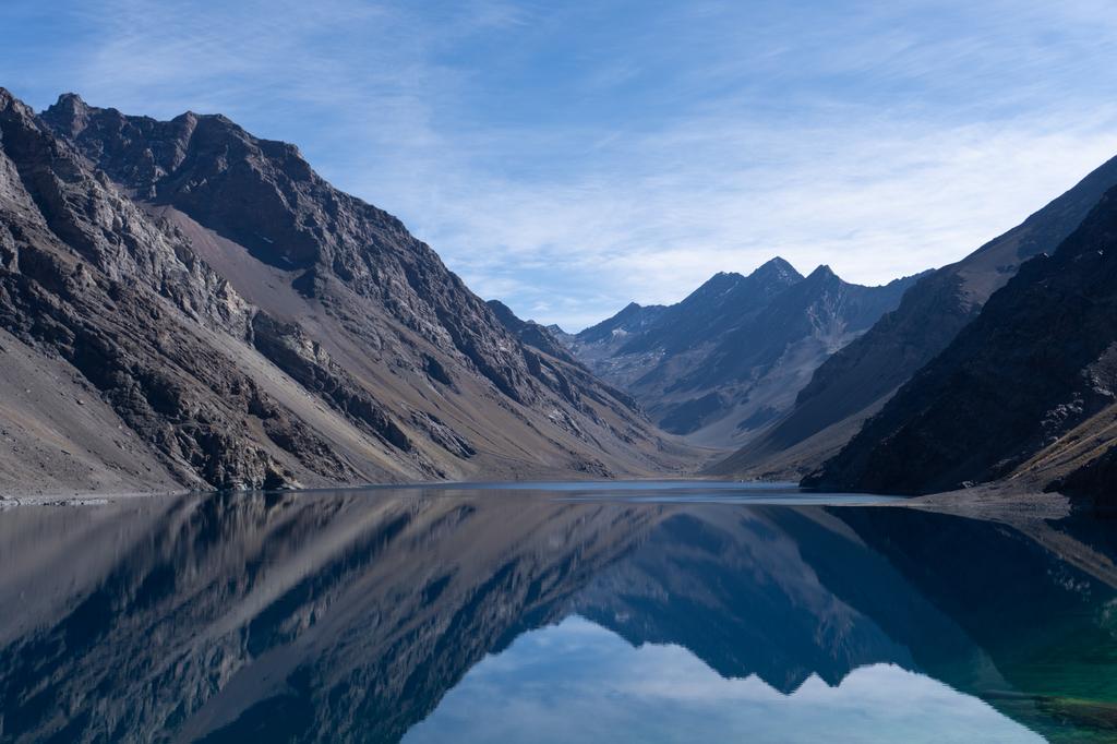 High-altitude mountain lake with mirror-like reflections of steep rocky peaks under blue sky in the Andes