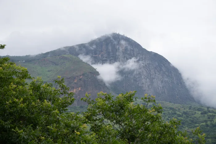 The rocky face of Ibituruna Peak partially shrouded in low clouds, rising above the green slopes and dense forest canopy.