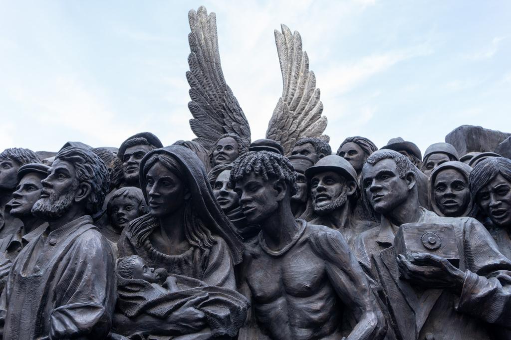 A large bronze sculpture depicting a dense crowd of diverse people, with a pair of angel wings rising above them against a bright sky.