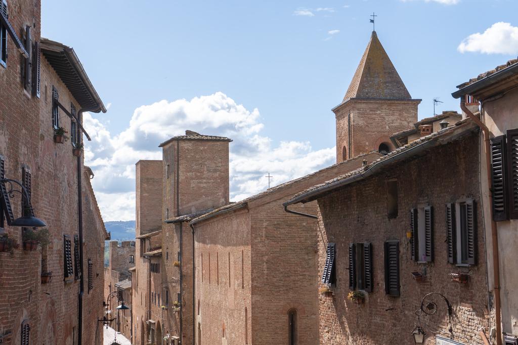 A street of tall medieval brick buildings in a Tuscan hilltop town, with shuttered windows and a pointed church tower rising above the rooftops against a blue cloudy sky.