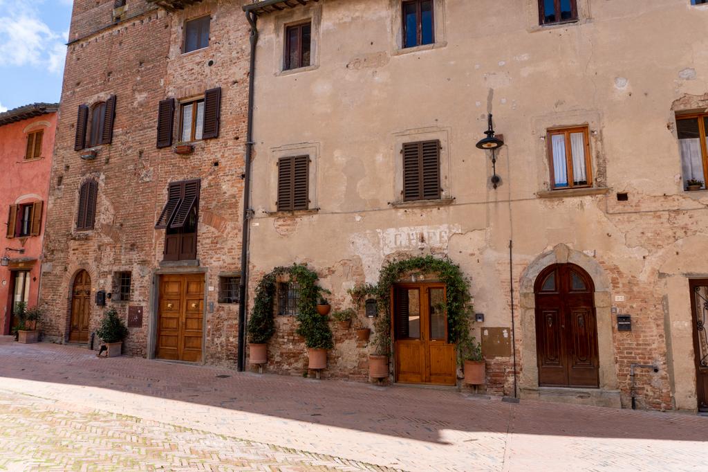 A sun-drenched medieval piazza in a Tuscan village, with terracotta and stone-coloured buildings, wooden doors, potted plants and climbing plants adorning the facades.