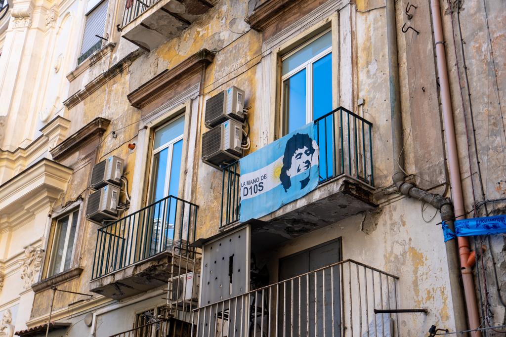 The weathered yellow facade of a Naples apartment building with a balcony displaying an Argentine flag with Maradona's face and the text 'La Mano de D10S'.