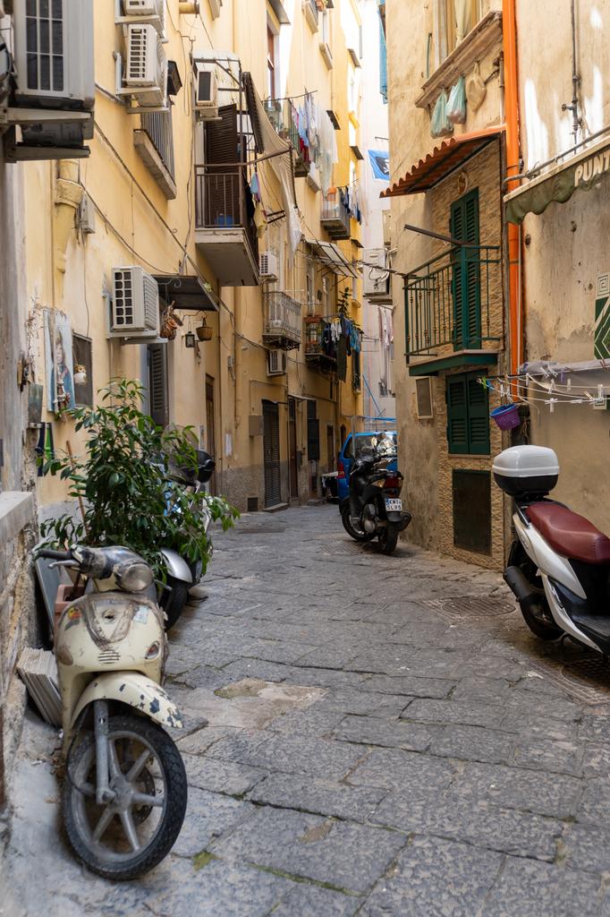 A narrow cobblestone alley in Naples with scooters and Vespas parked along the walls, laundry hanging from balconies above, and tall colourful buildings on either side.