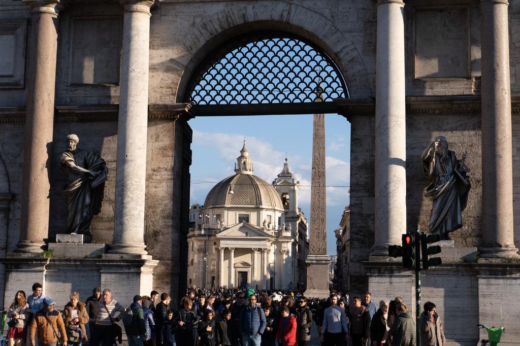 A crowd of people seen through the large arch of Porta del Popolo in Rome, with the twin churches and an Egyptian obelisk visible in Piazza del Popolo beyond.