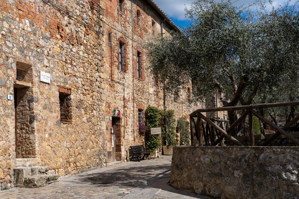 A rustic stone building in a small Italian village, with an old olive tree in a circular stone planter and a wooden fence in the foreground on a sunny day.