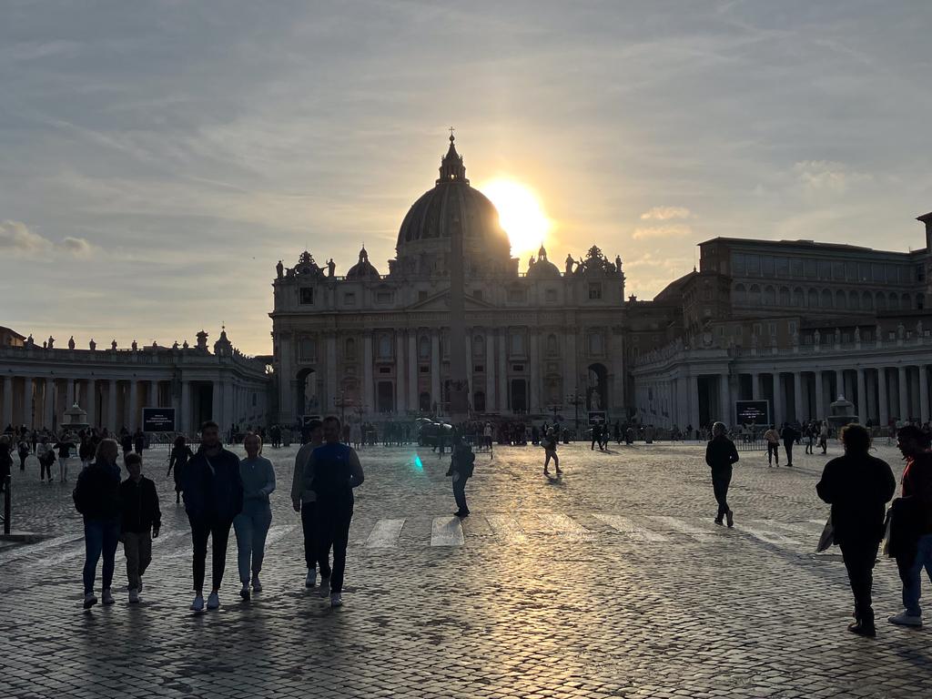 St. Peter's Basilica at Vatican City seen from St. Peter's Square at sunset, with the sun behind the dome casting a golden glow and silhouettes of visitors walking across the cobblestones.