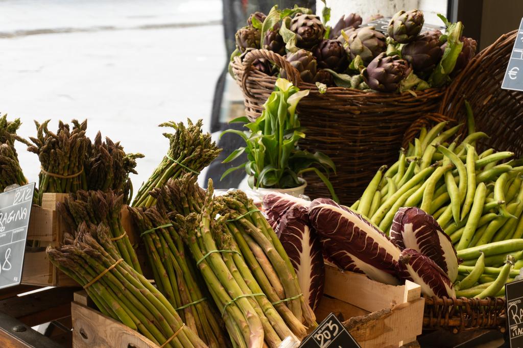 A colourful display of fresh vegetables at an outdoor market, including bundles of green asparagus, purple artichokes in a wicker basket, radicchio and green beans.