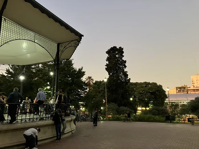 An ornate gazebo in a Belgrano park at dusk with people gathered around it, tall trees and city lights in the background under a pale pink sky.